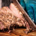 A gloved hand slicing juicy grilled beef brisket flat cut on a wooden cutting board, showing the pepper-crusted crust and pink interior