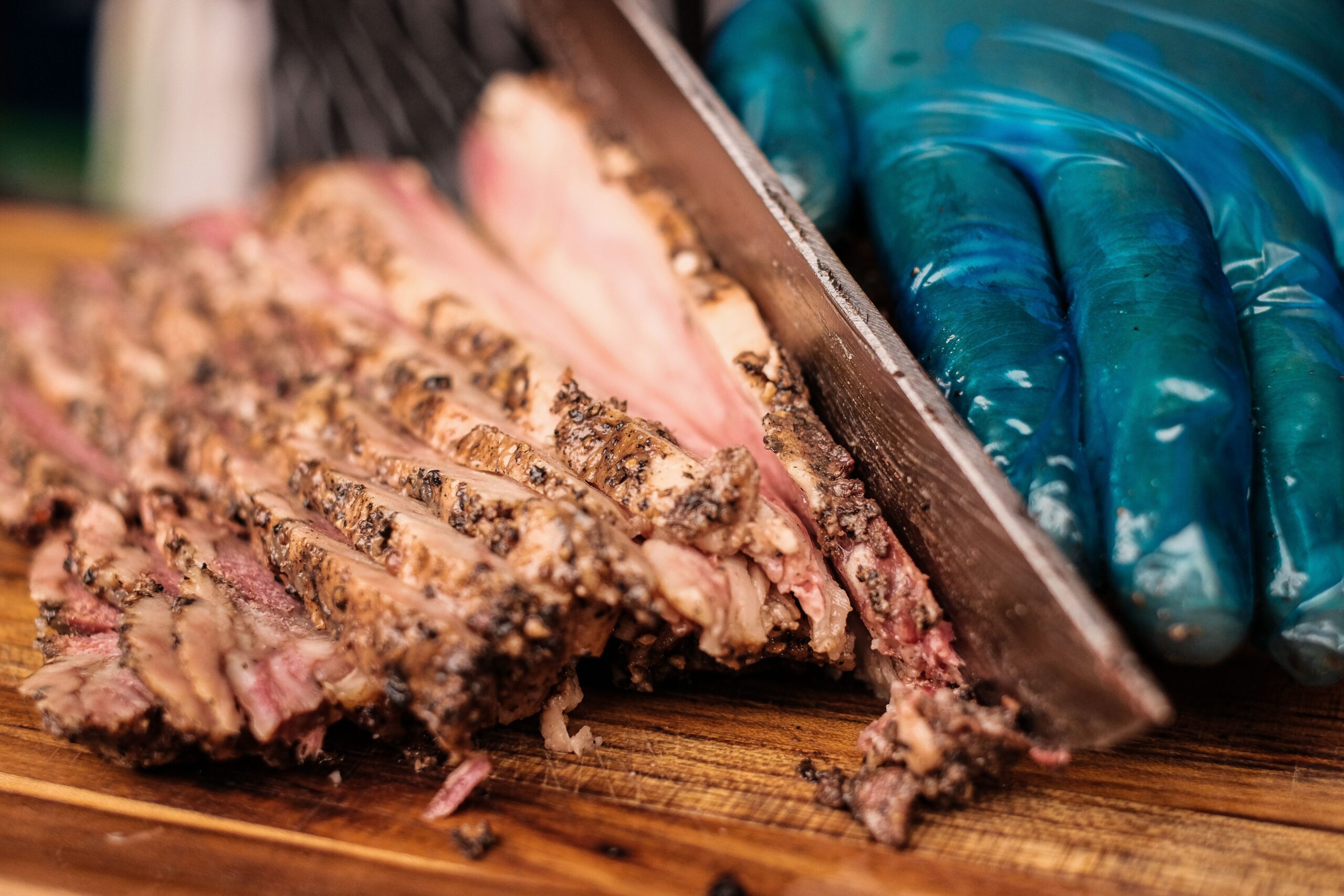 A gloved hand slicing juicy grilled beef brisket flat cut on a wooden cutting board, showing the pepper-crusted crust and pink interior