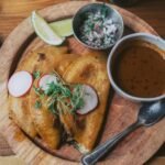 Crisp folded beef tacos on a plate with a cup of red consommé, radish and lime
