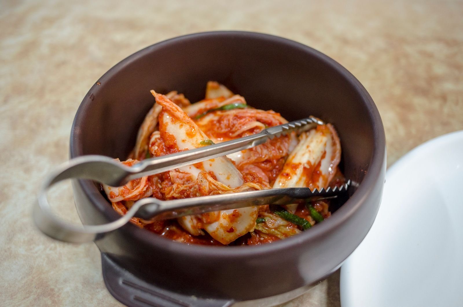 Napa cabbage kimchi in a dark ceramic bowl with metal tongs
