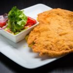 Golden crumbed schnitzel on a white plate beside a small bowl of fresh salad