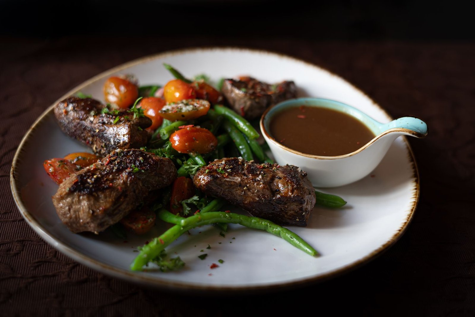 Sliced seared steak with green vegetables and a small jug of pepper cream sauce on a white plate