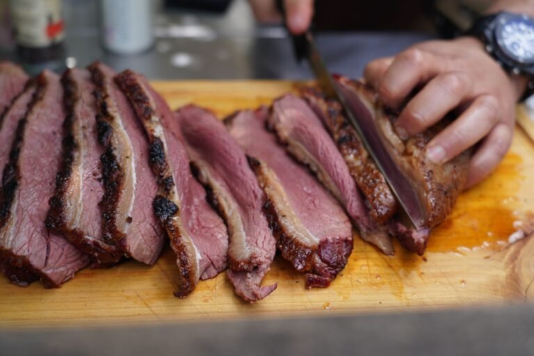 Hand slicing a pink, medium rare beef roast into thin slices on a wooden board