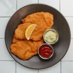 Two golden fried schnitzels with a lemon wedge on a dark plate, seen from above