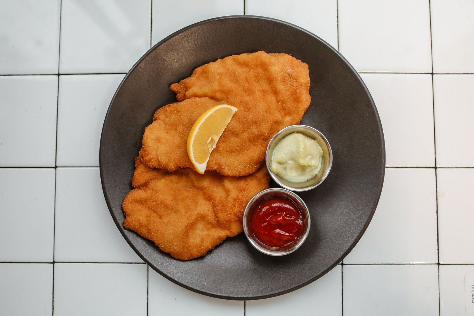 Two golden fried schnitzels with a lemon wedge on a dark plate, seen from above